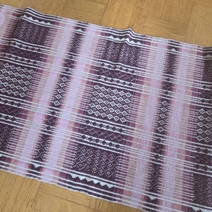 May include: A woven textile with a geometric pattern in shades of purple, pink, and white. The design features vertical stripes and diamond-shaped motifs, creating a textured appearance. The fabric is likely used for home decor or crafting.