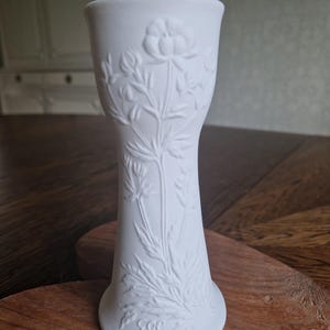 May include: A white, ceramic vase with a flared top and a detailed floral relief design. The vase features a slender, elongated shape and a textured surface. The floral pattern includes flowers and leaves. The base of the vase has a signature.