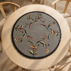 Op de afbeelding: Een ronde grijze placemat met een getextureerd oppervlak en een zwarte franjerand. Het midden is versierd met een geborduurd bloemenrankpatroon in tinten rood, geel, blauw en roze, gerangschikt in een symmetrisch cirkelvormig ontwerp.