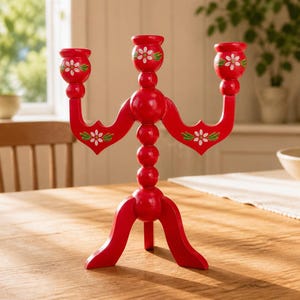 May include: A bright red wooden candelabra featuring three candle holders arranged in a tiered structure. The piece is decorated with hand-painted white daisy motifs with green leaves on the cups and arms, supported by a central column and tripod base.