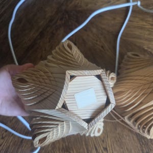 Może przedstawiać: Drewniana, geometryczna lampa wisząca o warstwowym wzorze. Oprawa wykonana jest z jasnego drewna i ma pięciokątną podstawę. Do oprawy przymocowane są białe przewody elektryczne.