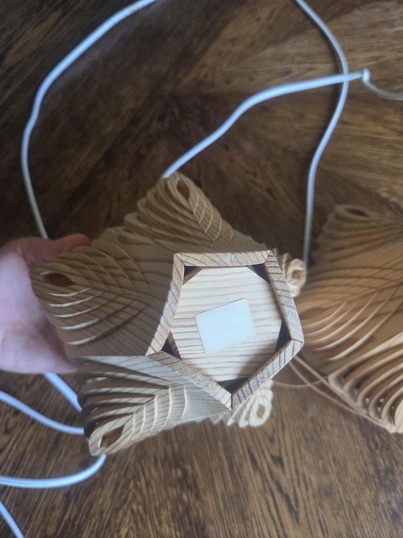 Może przedstawiać: Drewniana, geometryczna lampa wisząca o warstwowym wzorze. Oprawa wykonana jest z jasnego drewna i ma pięciokątną podstawę. Do oprawy przymocowane są białe przewody elektryczne.