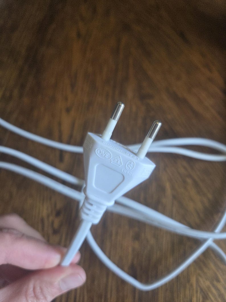 Może przedstawiać: Biała wtyczka elektryczna z dwoma bolcami i białym kablem. Wtyczka ma na swojej powierzchni nadrukowane kilka symboli bezpieczeństwa. Kabel jest biały i wydaje się być wykonany z tworzywa sztucznego. Tło to drewniana powierzchnia.