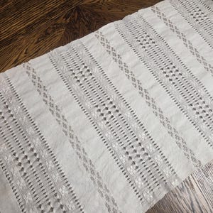 May include: A rectangular, off-white, open-weave textile with vertical stripes. The stripes alternate between solid and patterned sections, creating a textured appearance. The textile is likely a decorative piece, such as a table runner or a scarf.