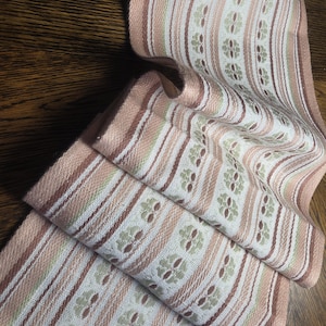 Puede incluir: Un textil plegado con un patrón floral repetitivo. La tela presenta rayas verticales en tonos de blanco, marrón y rosa. El diseño floral es de color verde apagado. Los bordes están acabados con un borde rosa. El textil es probablemente un camino de mesa o un paño decorativo.