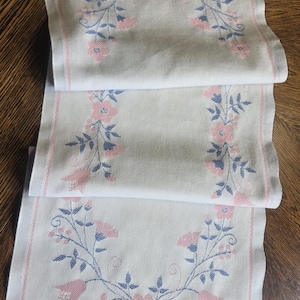 May include: White table runner with pink and blue floral embroidery. The runner has a repeating pattern of pink flowers and blue leaves, framed by a pink border. The ends have a fringe.