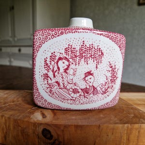 Puede incluir: Una botella rectangular de cerámica blanca con un patrón floral rojo y una escena decorativa de dos personas bajo un árbol. La botella tiene un pequeño tapón cilíndrico blanco y está sobre una superficie de madera.