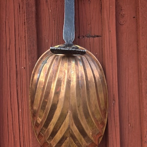 May include: A vintage, ornate brass-colored kitchen utensil, possibly a mold or decorative piece, hangs against a weathered red wooden wall. The utensil has a long, dark metal handle and a large, oval-shaped bowl with vertical ridges.