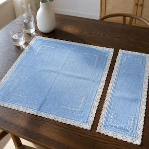 Op de afbeelding: Een set van een vierkante en een rechthoekige lichtblauwe placemat van stof, voorzien van een witte kanten rand en witte geborduurde details in de hoeken. De stof heeft een zichtbare weefstructuur en de kanten rand is fijn afgewerkt.