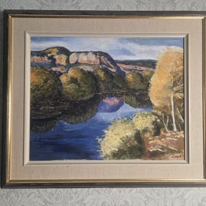 May include: A framed landscape oil painting depicting a dark blue river reflecting a mountain range under a cloudy sky. The scene features golden-yellow trees on the right bank and dark green foliage on the left, all enclosed in a wooden frame.