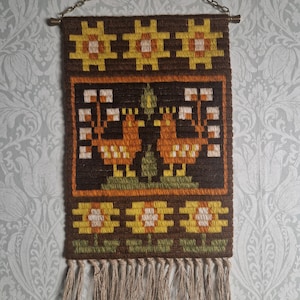 Puede incluir: Un tapiz de pared tejido con un fondo marrón y un patrón floral repetido en amarillo, naranja y blanco. El diseño central presenta dos pájaros estilizados. El borde inferior tiene una franja.