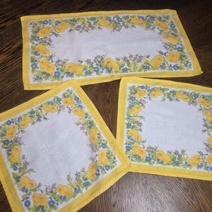 Op de afbeelding: Een set van drie vintage paaslinnen. De set bevat een rechthoekige tafelloper en twee vierkante placemats. Elk stuk heeft een witte kern met een rand van gele kuikens, blauwe bloemen en groen blad, omlijst door een gele rand.