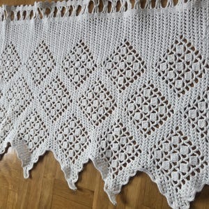 May include: A white, crocheted valance featuring a diamond pattern. The valance has a scalloped lower edge and a fringe along the top. The intricate openwork design is visible throughout, creating a delicate and airy appearance, suitable for a window.