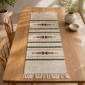Pode incluir: Um caminho de mesa retangular bege com textura tecida e extremidades com franjas. O design inclui listras horizontais em verde e laranja, acentuadas por padrões geométricos repetidos em preto, verde e laranja por todo o tecido.