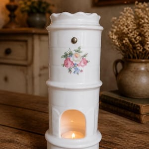 May include: A white ceramic oil burner with a cylindrical shape and a scalloped top edge. The exterior features a floral decal of pink and white roses with green leaves and a small metal stud. A tealight candle glows inside the arched opening.