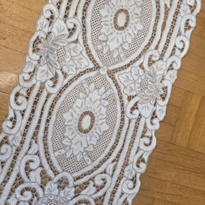 May include: A white, intricately designed lace table runner. The runner features floral and geometric patterns with a delicate, openwork design. The runner is placed on a wooden surface.