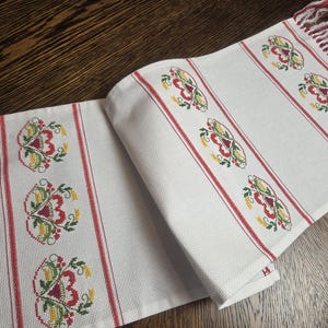 May include: White table runner with red stripes and embroidered floral patterns in red, yellow, and green. The runner has fringed edges in red and white, adding a decorative touch to the textile.