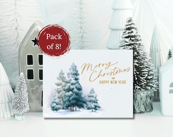 Può includere: Un pacco di otto biglietti di Natale con una scena di foresta invernale. I biglietti presentano un acquerello di alberi sempreverdi coperti di neve e le parole "Merry Christmas and Happy New Year" in oro. Case e alberi decorativi in ceramica bianca circondano i biglietti.