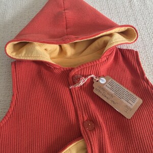 May include: A red hooded vest with a yellow flannel lining. The vest has a button closure and a pocket on the front. The tag reads "cotton outer, cotton flannel inner, size 1, £50."