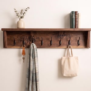 May include: A dark brown wooden shelf with eight black metal hooks. A beige tote bag, a patterned scarf, and a leather keychain with keys hang from the hooks. A small vase with greenery and three books sit on the shelf.