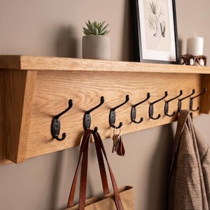 May include: A wooden wall-mounted shelf with eight black metal hooks. A brown leather tote bag and keys hang from the hooks. A framed picture, a candle, and a small potted plant sit on the shelf above.