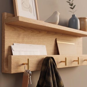 May include: A light oak wood wall shelf with a top shelf holding a framed picture, a white bird figurine, and small vases. The lower section has a mail slot, brass hooks, and a hanging scarf. Keys hang from a brass hook.