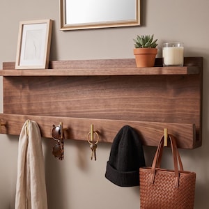 May include: A dark wood wall shelf with a top shelf and a lower shelf with brass hooks. Items on the shelf include a framed picture, a small potted plant, and a candle. Below the shelf hang a scarf, sunglasses, keys, a beanie, and a brown tote bag.
