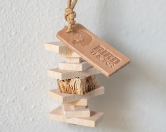 Può includere: Un giocattolo di legno per uccelli con un'etichetta in pelle che dice "Bird Tricks". Il giocattolo è fatto di blocchi di legno impilati e un pezzo di legno naturale. Il giocattolo è appeso a una corda marrone.