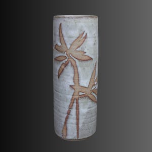 May include: A cylindrical ceramic vase with a speckled off-white glaze and a textured surface. The exterior features a hand-painted brown botanical design depicting bamboo-like stalks and leaves that wrap around the vertical body of the vessel.
