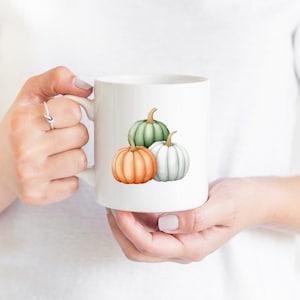 Op de afbeelding: Witte keramische mok met een ontwerp van drie pompoenen. De pompoenen zijn groen, wit en oranje.