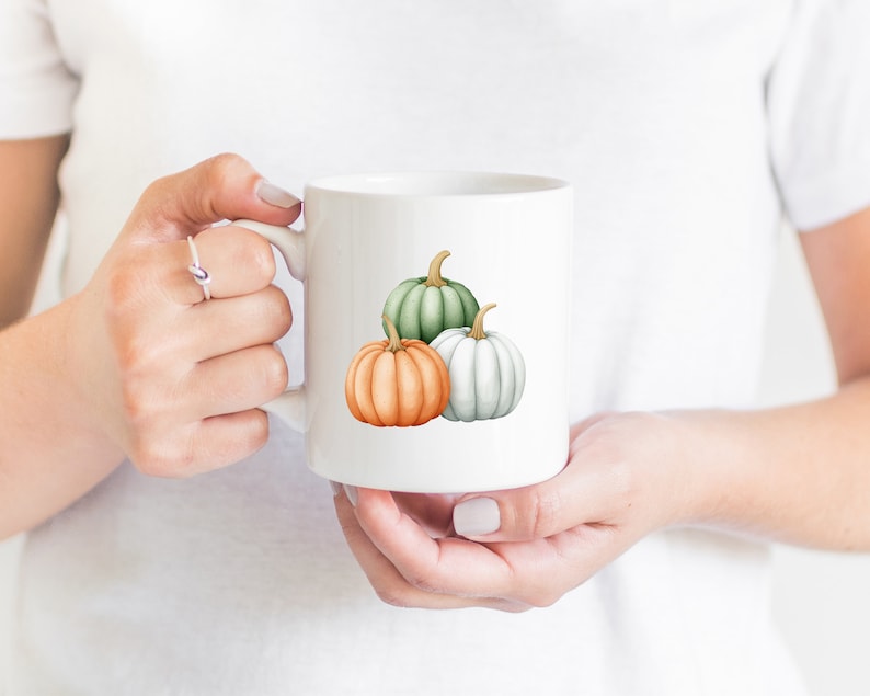 Op de afbeelding: Witte keramische mok met een ontwerp van drie pompoenen. De pompoenen zijn groen, wit en oranje.