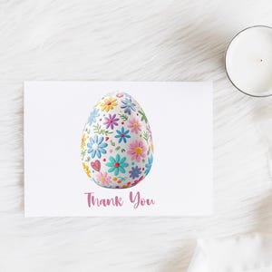 May include: A white greeting card with a floral Easter egg design and the words "Thank You" in pink script. The egg is covered in colorful flowers. A lit candle sits to the right.