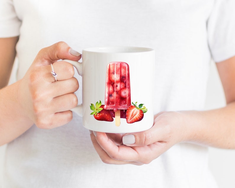 May include: White ceramic mug with a red and white strawberry popsicle design. The popsicle is filled with red fruit and has a wooden stick. There are two strawberries on either side of the popsicle.