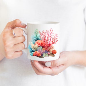 K&ouml;nnte beinhalten: Wei&szlig;e Keramiktasse mit einer bunten Illustration von Korallen und Muscheln. Die Korallen sind rot, rosa und orange, und die Muscheln sind wei&szlig;, blau und rosa.