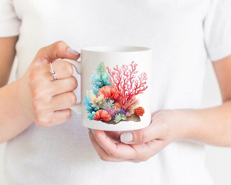 K&ouml;nnte beinhalten: Wei&szlig;e Keramiktasse mit einer bunten Illustration von Korallen und Muscheln. Die Korallen sind rot, rosa und orange, und die Muscheln sind wei&szlig;, blau und rosa.