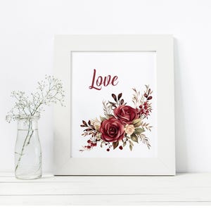 May include: A framed print with the word "Love" in burgundy script. The print features a floral arrangement of burgundy and cream roses with dark green and brown leaves. A small glass vase with white flowers is to the left.