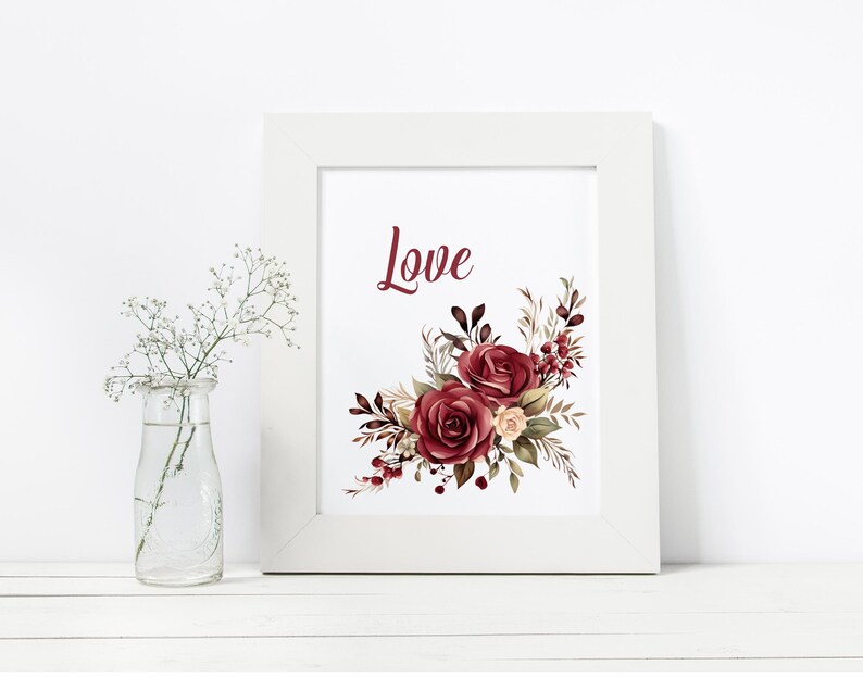 May include: A framed print with the word "Love" in burgundy script. The print features a floral arrangement of burgundy and cream roses with dark green and brown leaves. A small glass vase with white flowers is to the left.