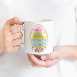 May include: White ceramic mug featuring a colorful Easter egg design. The egg is decorated with pink, yellow, and blue sections, adorned with floral and geometric patterns. The mug is held by a person.