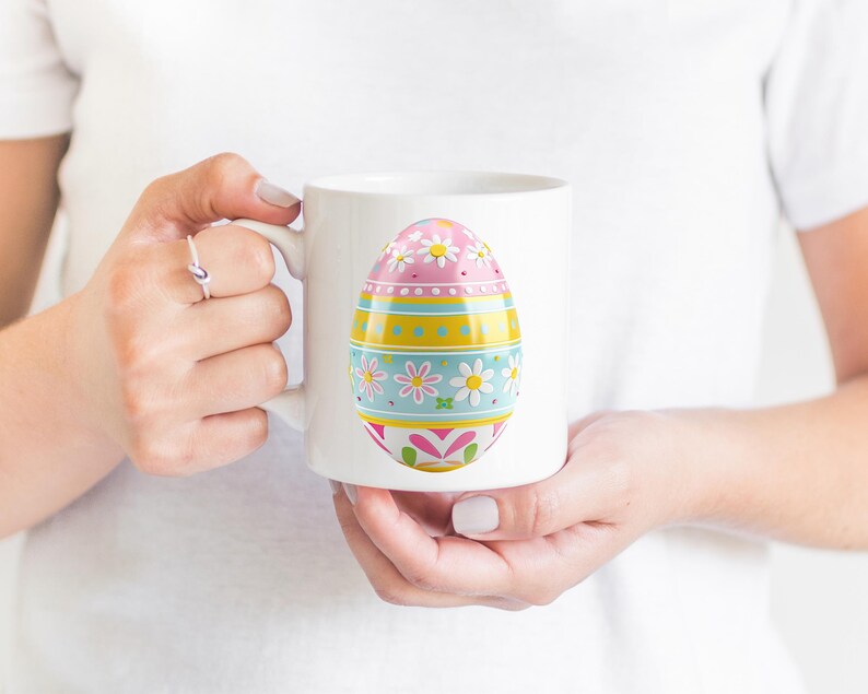 May include: White ceramic mug featuring a colorful Easter egg design. The egg is decorated with pink, yellow, and blue sections, adorned with floral and geometric patterns. The mug is held by a person.