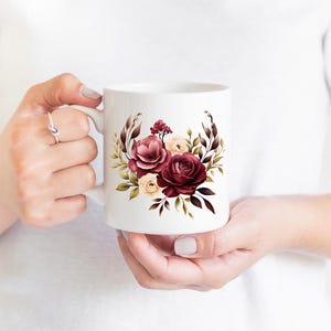 May include: White ceramic mug featuring a floral design with burgundy, cream, and green accents. The flowers are arranged in a circular pattern. The mug has a standard handle and is held by a person.