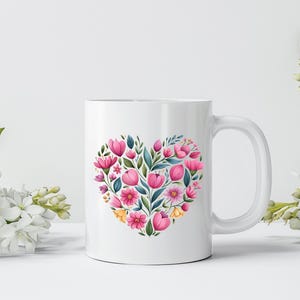 May include: White ceramic mug featuring a heart-shaped floral design. The heart is composed of pink tulips, other pink and yellow flowers, and green leaves. The mug has a white handle and is set against a white background.