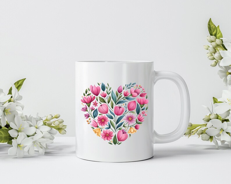 May include: White ceramic mug featuring a heart-shaped floral design. The heart is composed of pink tulips, other pink and yellow flowers, and green leaves. The mug has a white handle and is set against a white background.