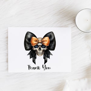 Pode incluir: Uma caveira branca com um la&ccedil;o preto e laranja. A caveira tem olhos pretos e um nariz preto. O la&ccedil;o est&aacute; amarrado em volta da cabe&ccedil;a da caveira. O texto "Thank You" est&aacute; impresso abaixo da caveira.