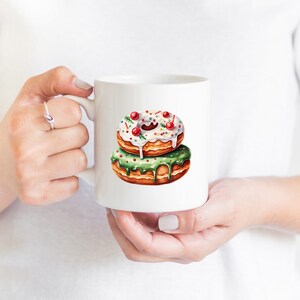 May include: White ceramic mug with a green and white donut with red sprinkles. The donut is stacked on top of another donut with white frosting and red sprinkles.