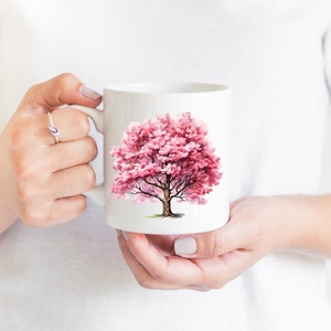 Pu&ograve; includere: Tazza in ceramica bianca con un'illustrazione ad acquerello rosa di un albero in fiore. L'albero ha un tronco spesso e rami con fiori rosa.