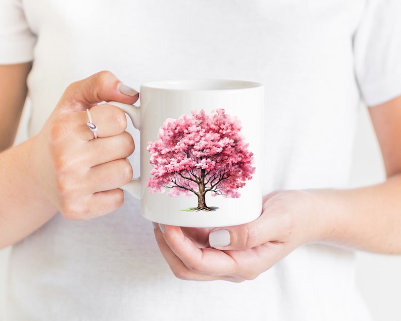 Pu&ograve; includere: Tazza in ceramica bianca con un'illustrazione ad acquerello rosa di un albero in fiore. L'albero ha un tronco spesso e rami con fiori rosa.