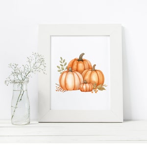 Op de afbeelding: Een wit frame met een aquarel van drie oranje pompoenen met groene bladeren en stelen. De pompoenen zijn gestapeld met de grootste pompoen onderaan en de kleinste pompoen bovenop.