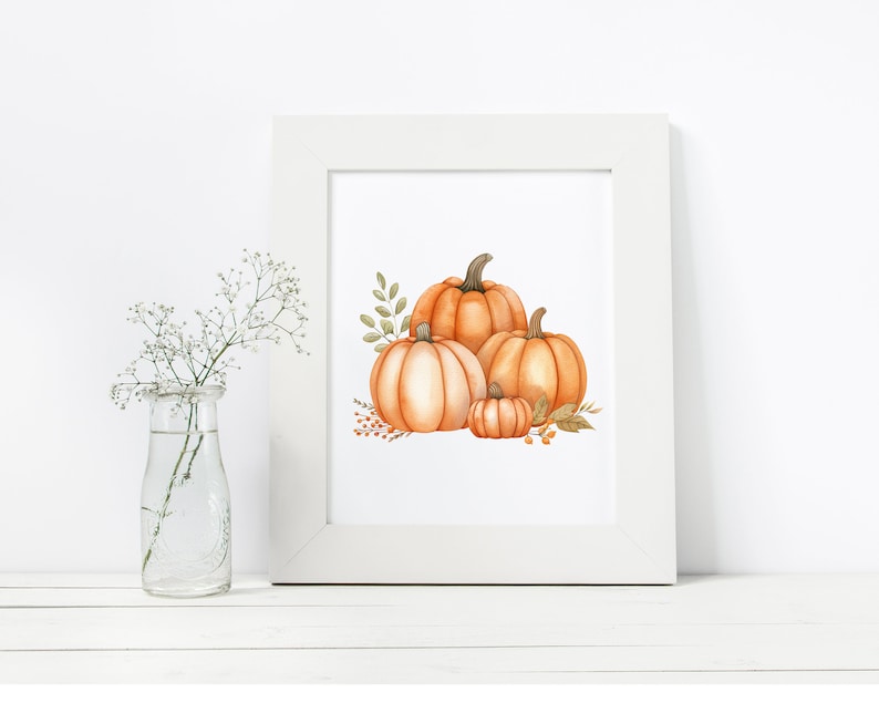 Op de afbeelding: Een wit frame met een aquarel van drie oranje pompoenen met groene bladeren en stelen. De pompoenen zijn gestapeld met de grootste pompoen onderaan en de kleinste pompoen bovenop.