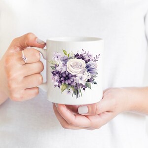 May include: White ceramic mug with a floral design of purple and white flowers. The bouquet includes roses, hydrangeas, and other blooms. The mug is held by a person.
