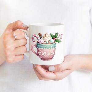May include: White ceramic mug featuring a watercolor illustration of a hot chocolate drink. The mug is decorated with a pink and teal design and filled with whipped cream, marshmallows, and a candy cane. A festive holiday theme.